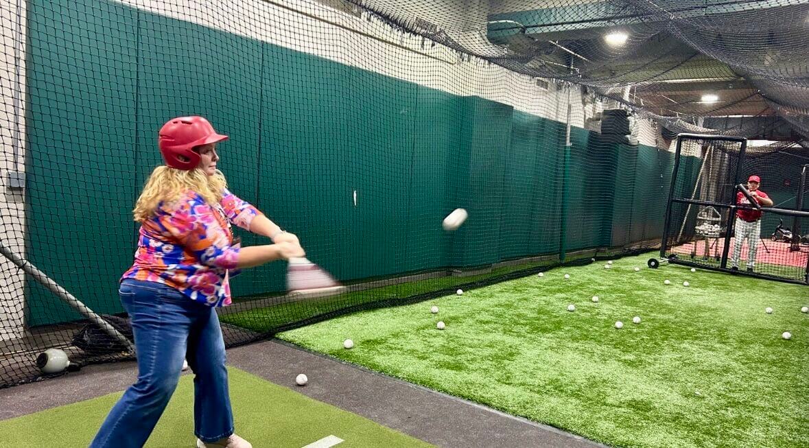 Woman hitting a ball inside a batting cage. Pitcher is in the background watching her.