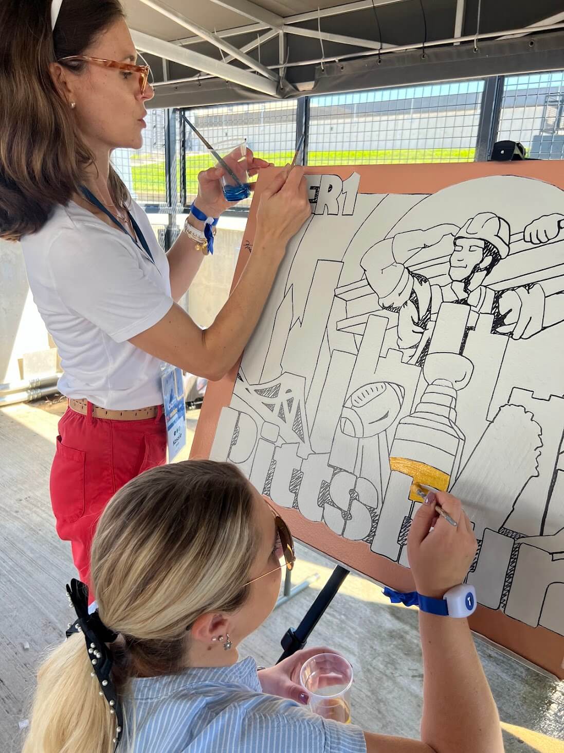 Two women painting a canvas depicting an image for Pittsburgh.