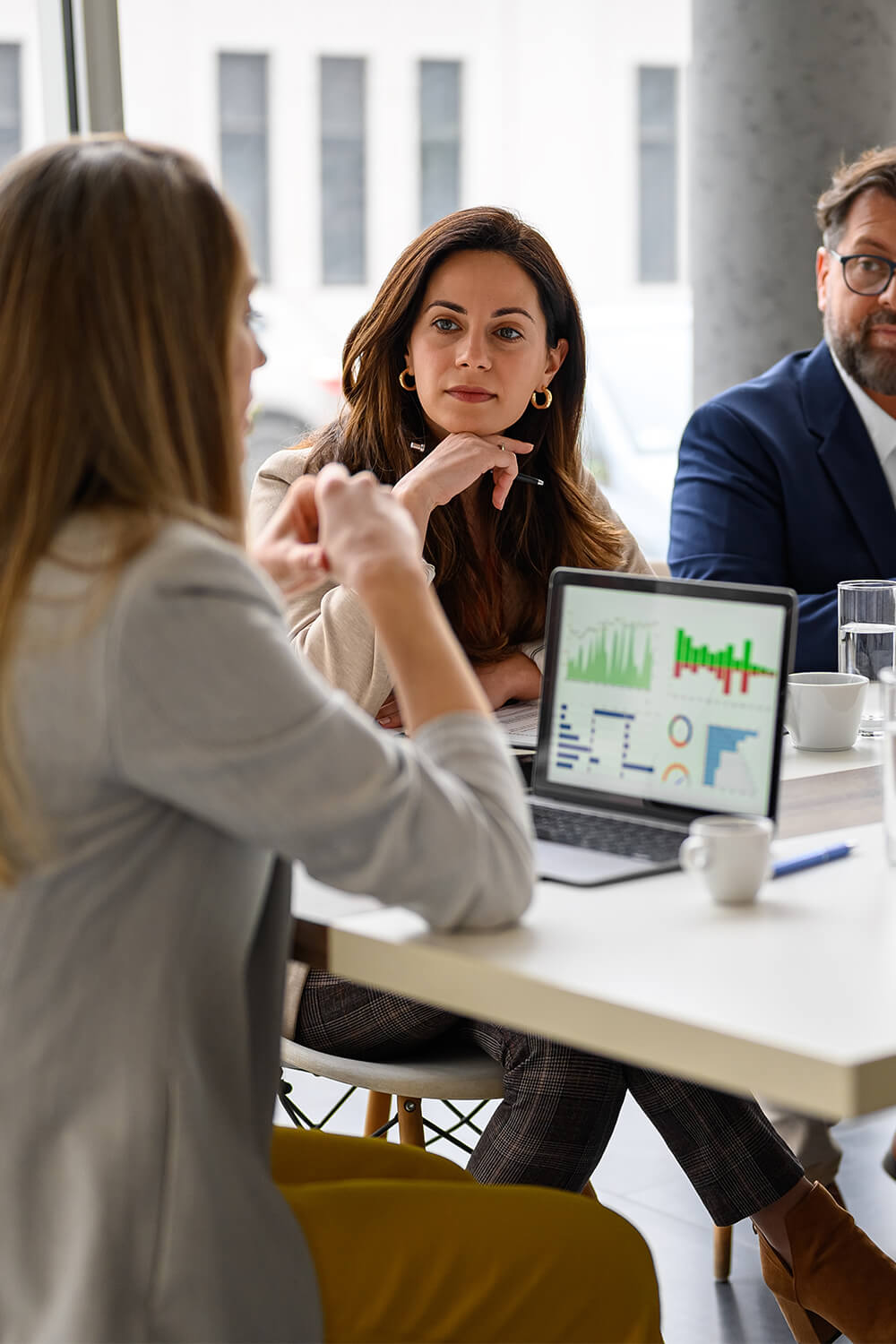 Four colleagues meet around a table reviewing charts and data on laptops during a business discussion.