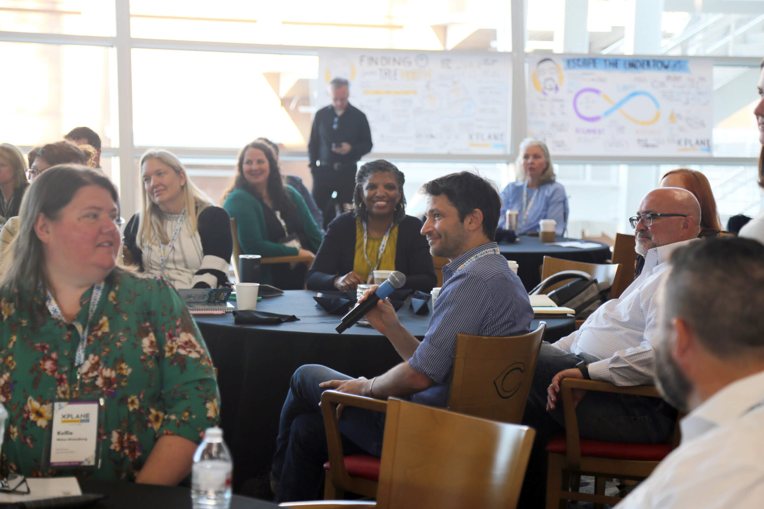 A man holding a microphone while surrounded by other people sitting at tables at a conference.