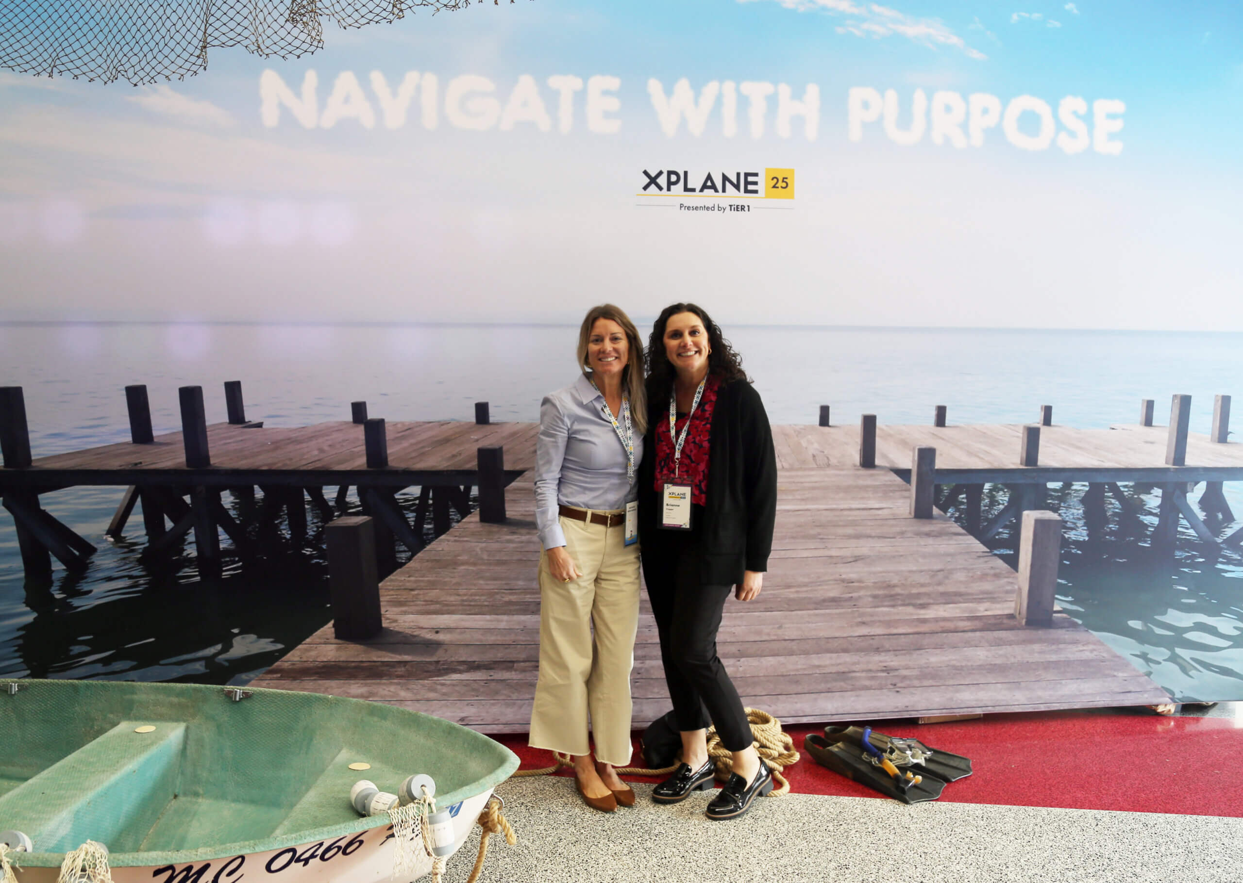 Two women standing in front of a backdrop depicting a dock for the conference theme Navigate with Purpose. There is a boat in the foreground of the photo.