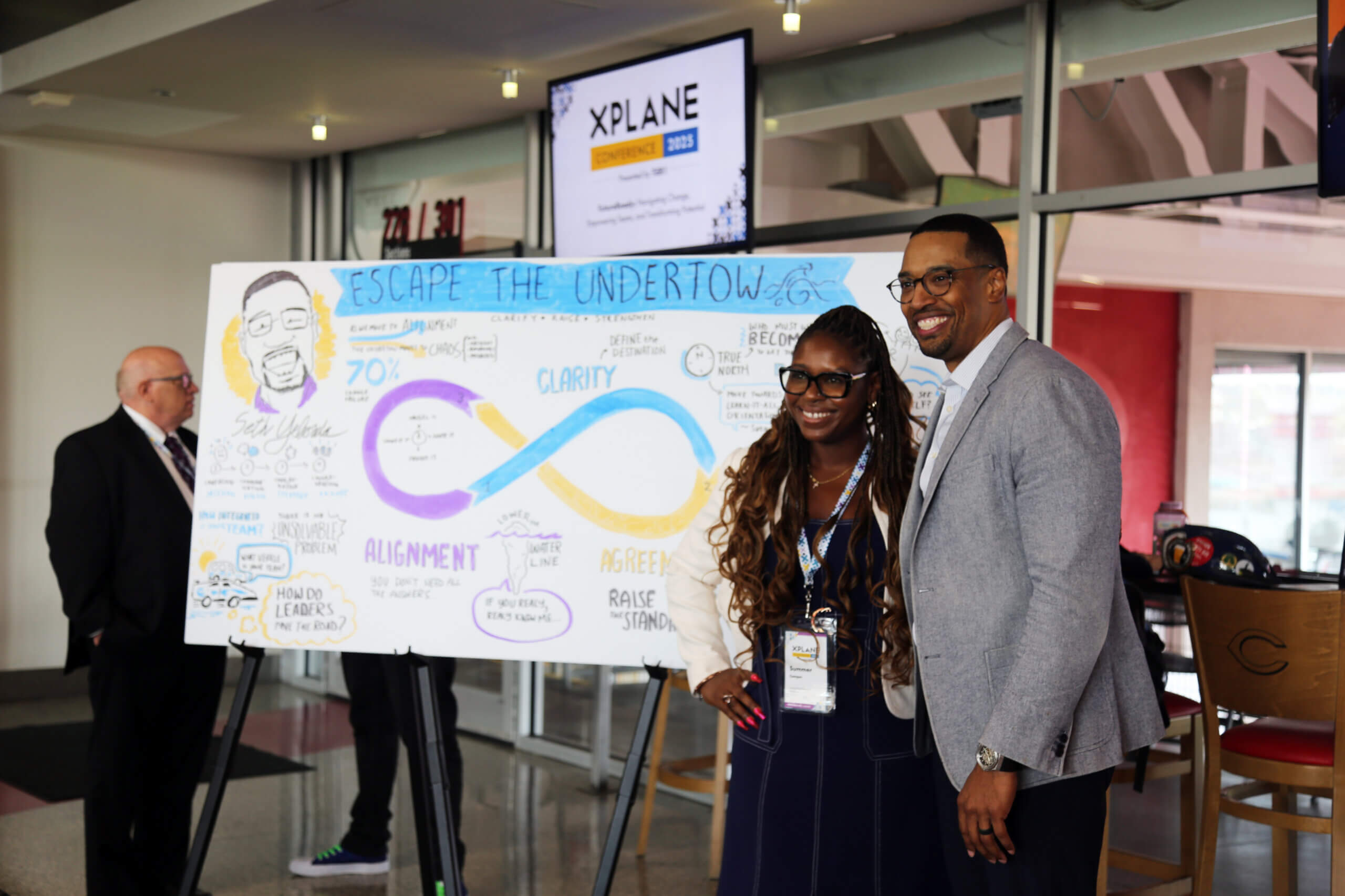 A man and a woman pose in front of a graphic facilitation depicting the man's keynote presentation.