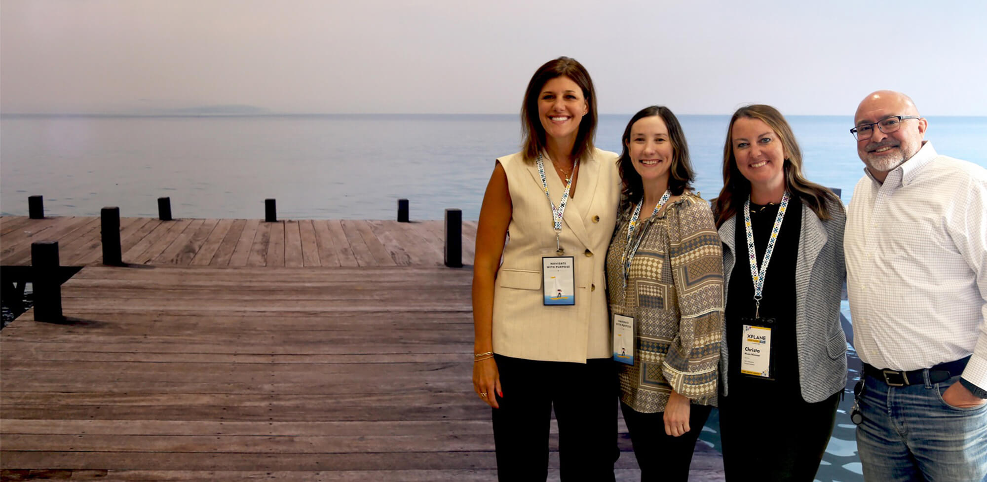Four colleagues wearing event badges stand together in front of a waterfront backdrop.