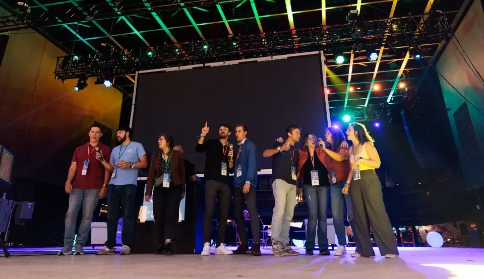 Group of colleagues stand on a stage together at an event, wearing badges and singing into microphones.