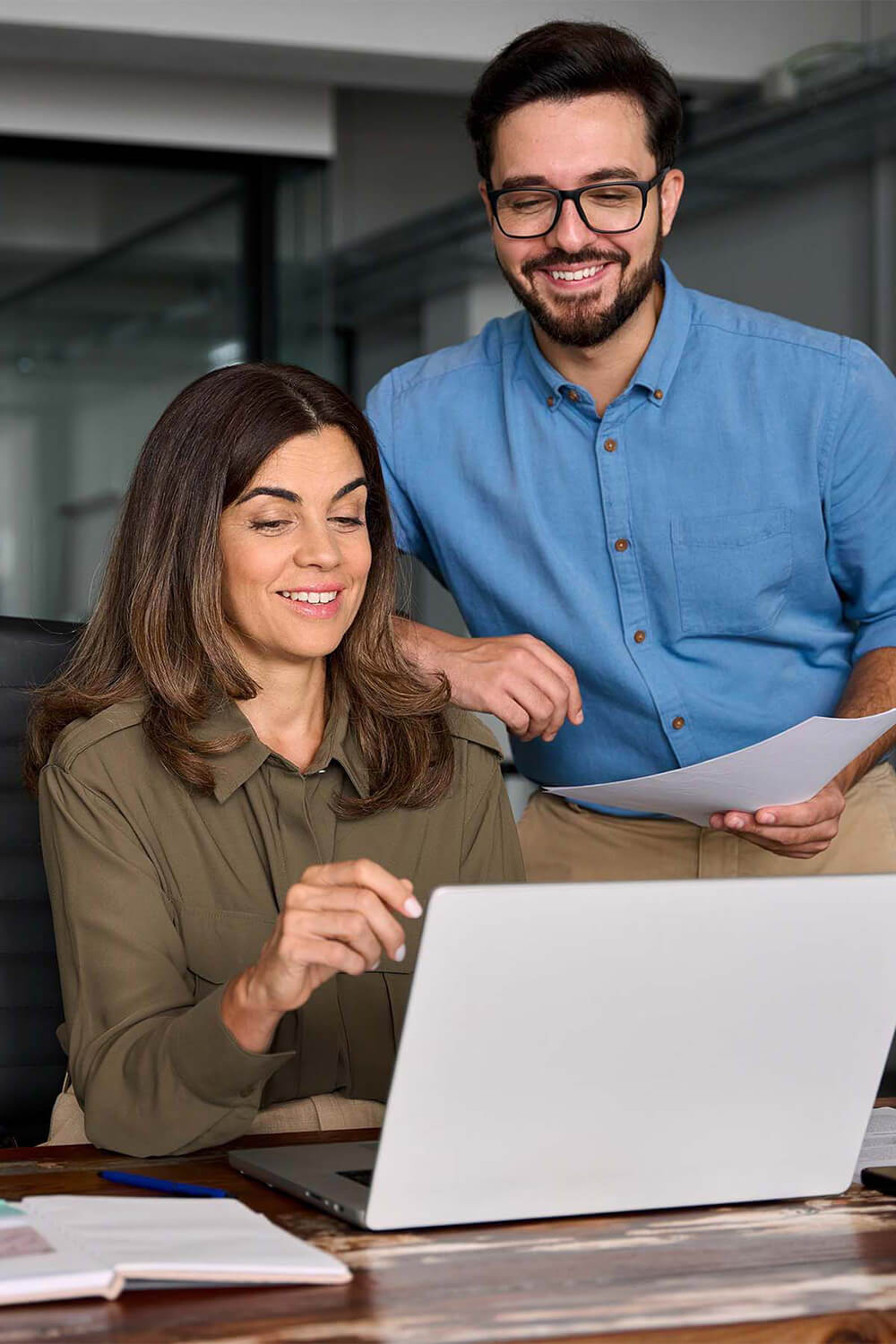 Two colleagues collaborate at a desk, reviewing a laptop and document together.